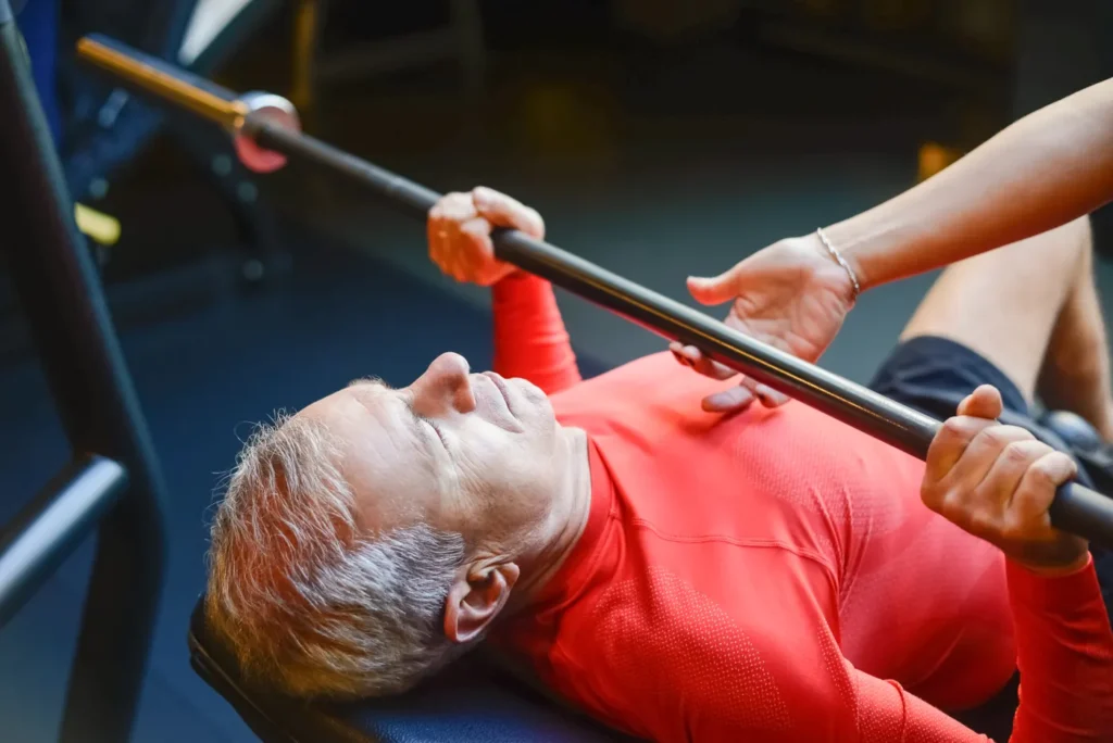 Older man performing a supervised barbell bench press in a gym