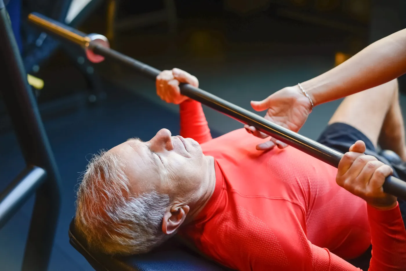Older man performing a supervised barbell bench press in a gym