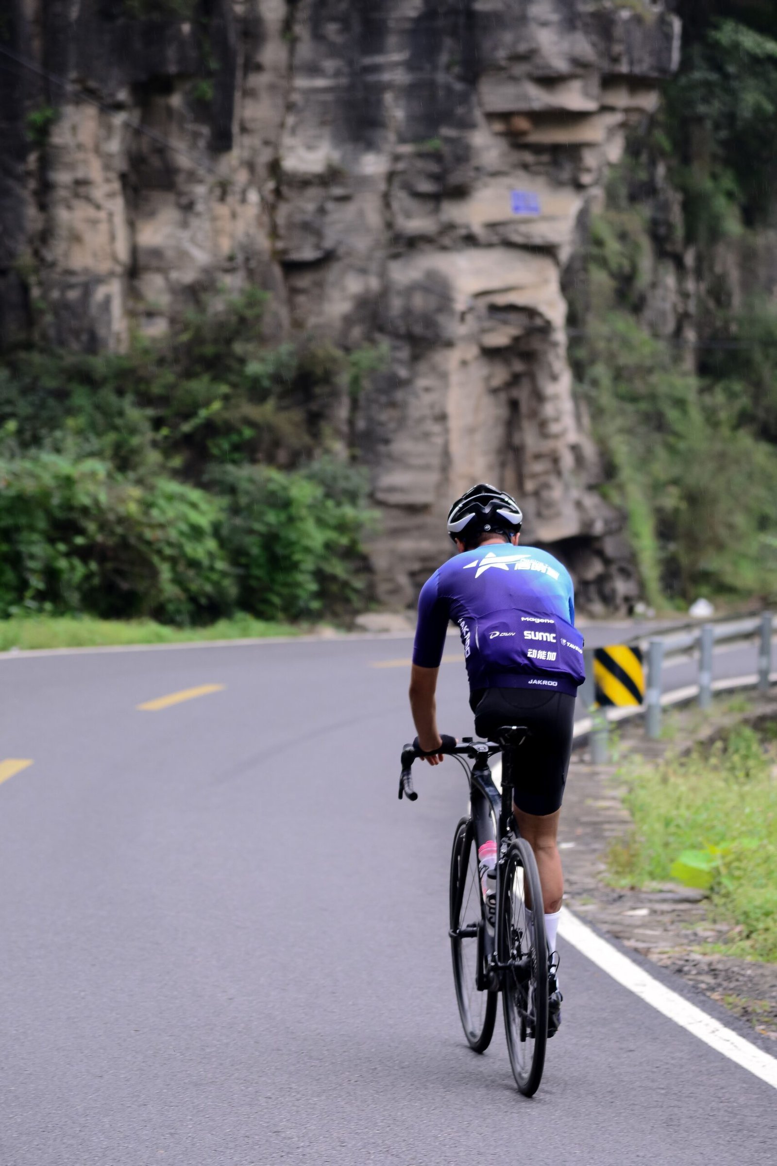 Cyclist in a blue and purple jersey riding a road bike beside rocky cliffs on a winding road