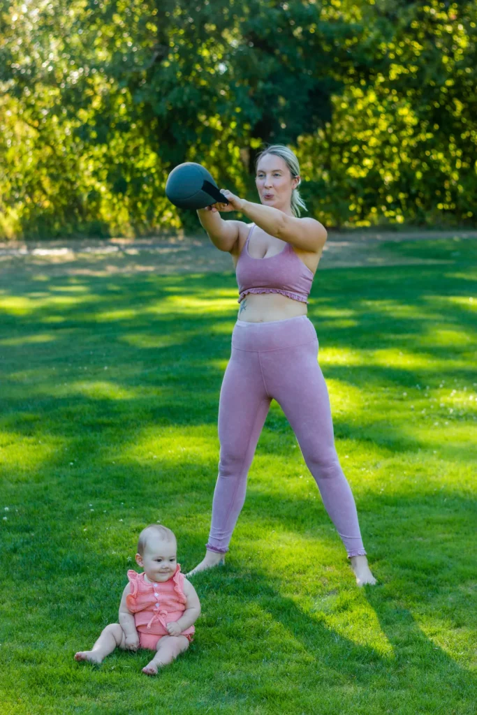 Mother lifting a kettlebell outdoors with her baby nearby